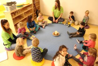 Erzieher sitzen mit Kindern in einem Kreis auf dem Boden und lachen