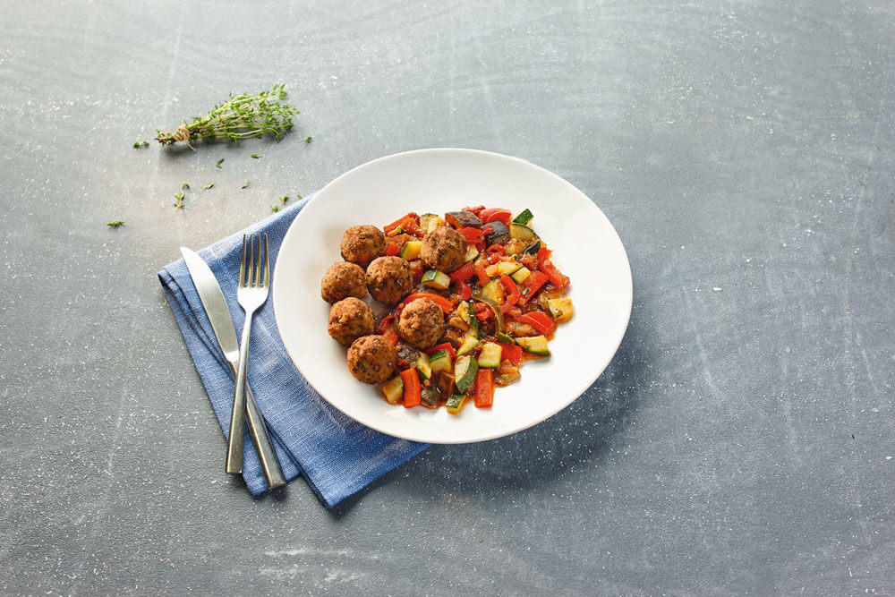 Auf einem Teller liegen Gemüsebällchen mit Zucchini-Paprika-Tomaten-Gemüse, ein Gericht vom apetito Menüservice.