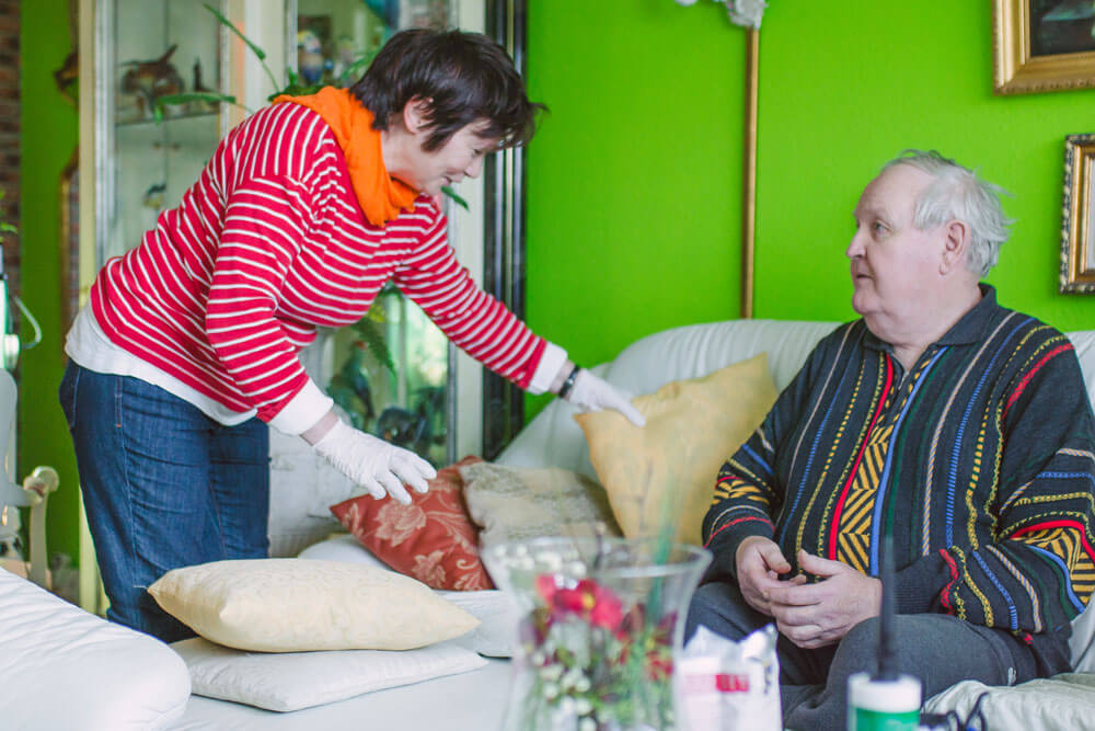 Putzkraft schüttelt ein Kissen für einen Senior auf, der auf dem Sofa sitzt.