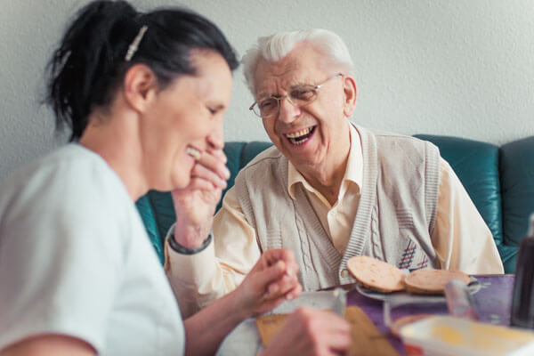 Pflegerin und Senior sitzen lachend am Esstisch.