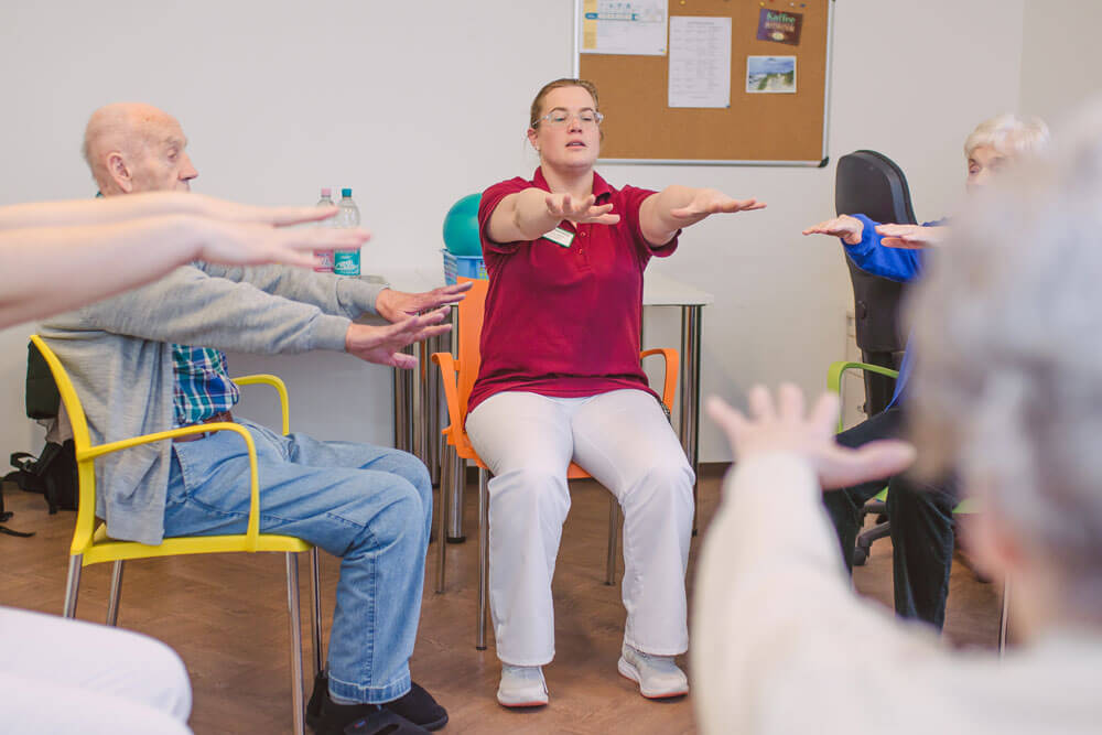 Senioren und Pfleger sitzen im Kreis und strecken gemeinsam ihre Hände nach vorne.