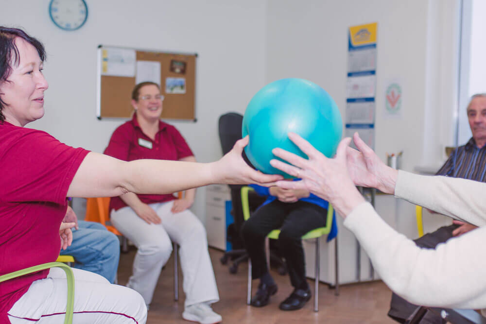 Pflegerinnen und Senioren reichen sich im Kreis einen Ball spielerisch weiter.