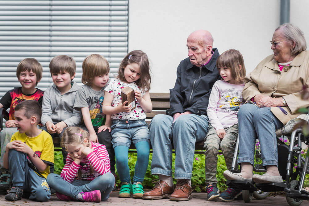 Senioren und Kinder sitzen auf einer Bank und schauen einem Kind gespannt zu, wie es etwas öffnet.
