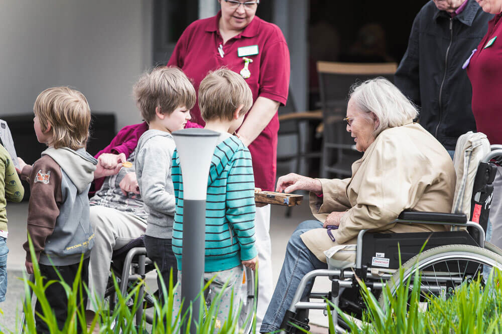 Kinder reichen einem Senior im Rollstuhl Kekse, während Pfleger, andere Senioren und Kinder um sie herum stehen.