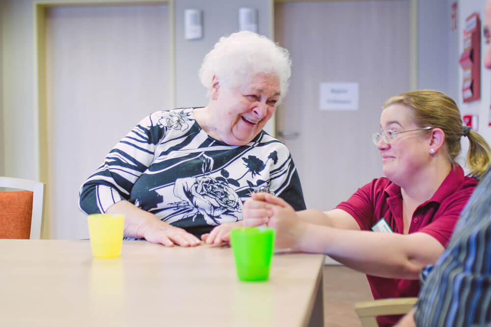 Eine Seniorin und eine Pflegerin lachen gemeinsam am Tisch.