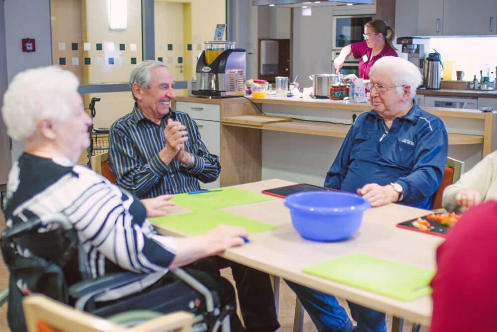 Lachende Senioren sitzen an einem Esstisch und haben Schneidebretter vor sich stehen.